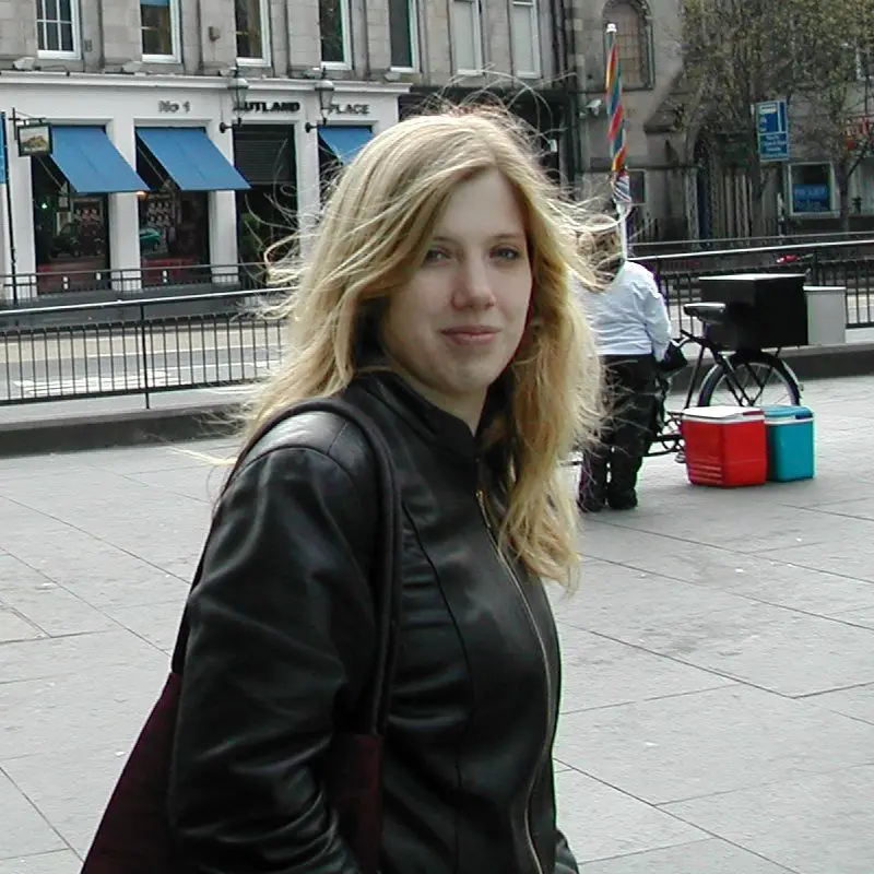 Sarah c.2004 twenty-something white woman with long blonde hair and wearing a black leather jacket, semi-smiles to camera as she’s walking along the street, slightly caught off guard by the unexpected photo.